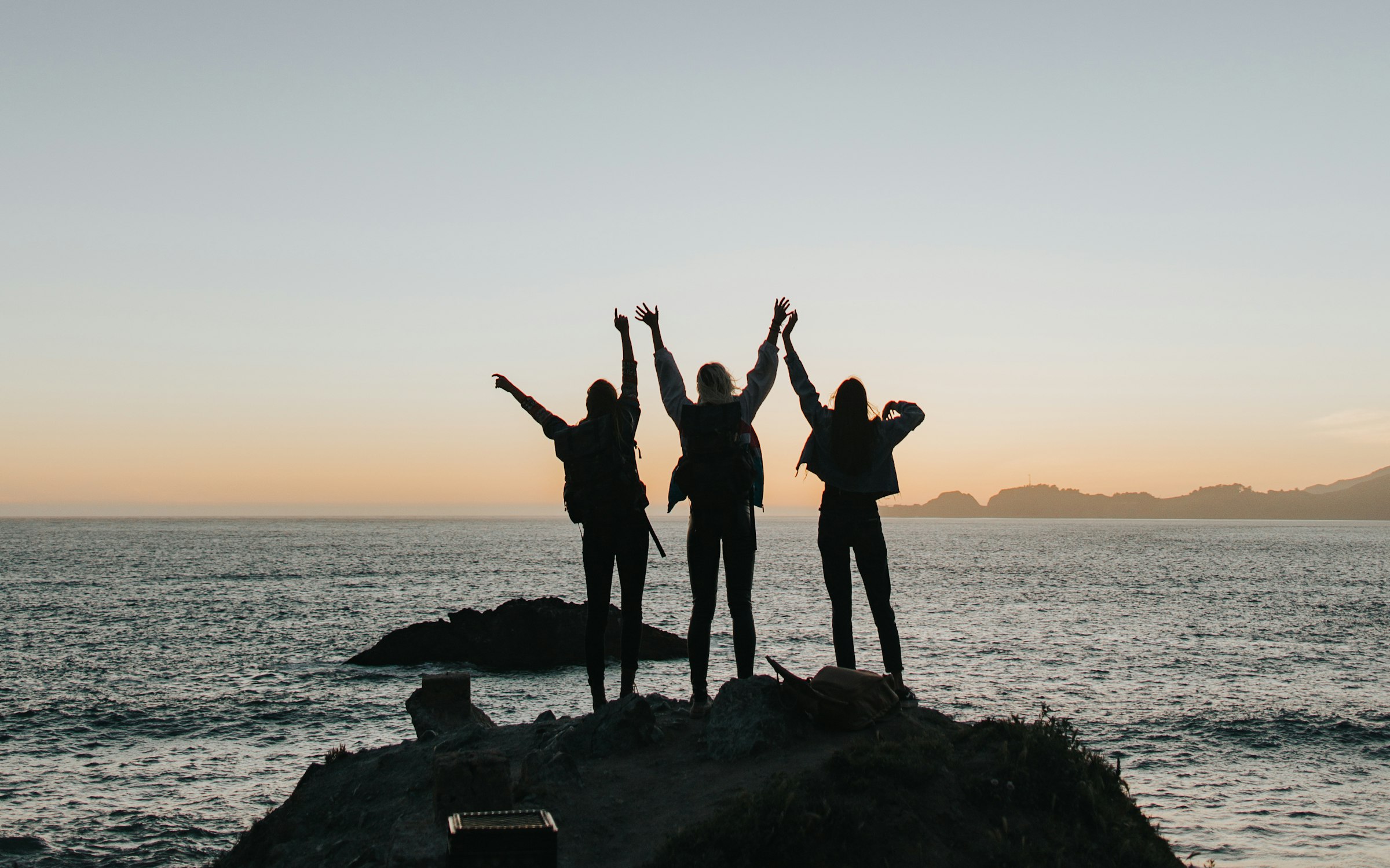 Three women cheering with arms raised on a cliff overlooking the ocean at sunset